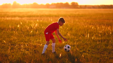 Junge-Junior-In-Einem-Roten-T-Shirt-Und-Turnschuhen-Bei-Sonnenuntergang,-Der-Mit-Einem-Fußball-Trainiert-Und-Sich-Darauf-Vorbereitet,-Ein-Fußballspieler-Zu-Werden.-Der-Weg-Zum-Traum.-Training-Bei-Sonnenuntergang-Und-Morgengrauen